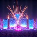 Panoramic view of thousands of people under a massive, colorful, carnival-like structure, illuminated by bright lights and fireworks overhead at night.